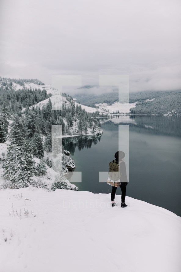 a woman standing on the edge of a snowy cliff looking down at a lake 