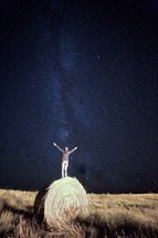 a man with raised hands standing on a hay bale under stars in the night sky 