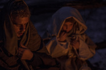 A man and child wrapped in Biblical clothing, sitting together near a fire.