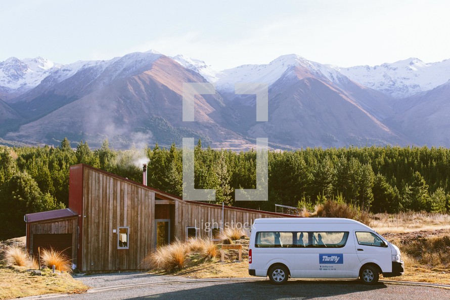 A white van parked in front of a log house at the base of a mountain range.