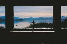 A cross on a windowsill looking out on a lake and mountain range.