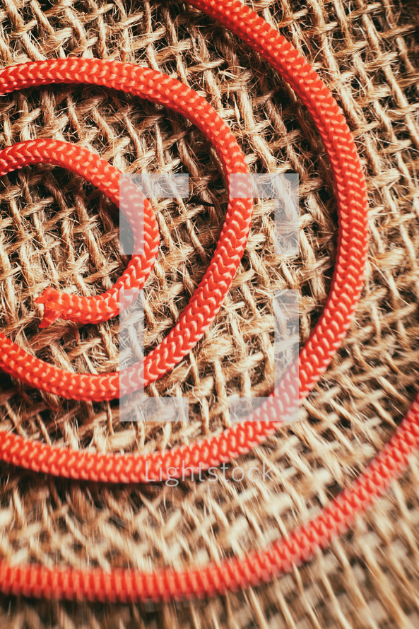 Red rope curled up forming a spiral on a background of light brown jute, with a blurred effect on the bottom