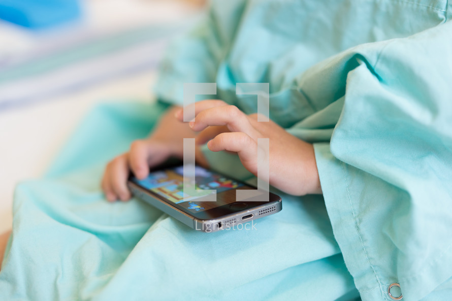 a boy child in a hospital gown playing games on a cellphone 