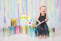 A laughing boy surrounded by colorful paint cans and Easter eggs.