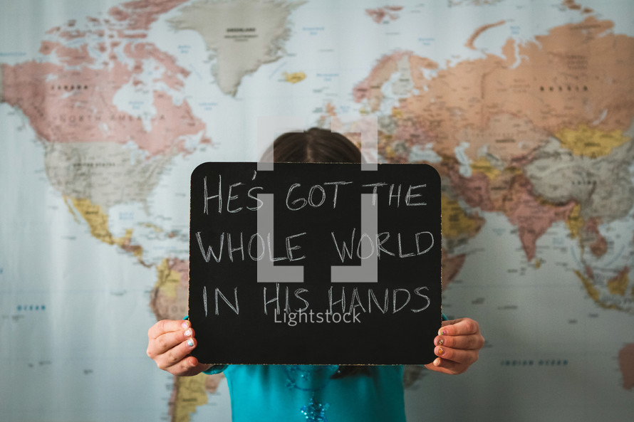 A girl in front of a world map holding a sign - He's got the whole world in his hands 