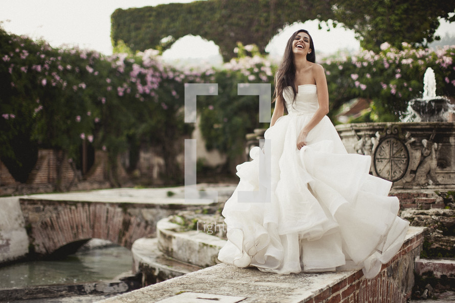 A smiling bride poses in her wedding dress near a fountain.