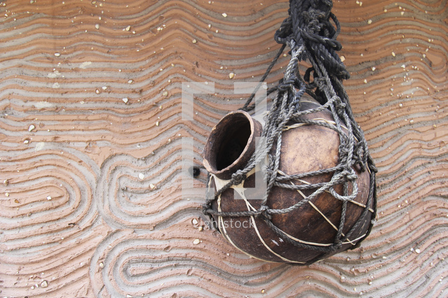 Clay water jug in rope holder hanging on decorative clay wall. 