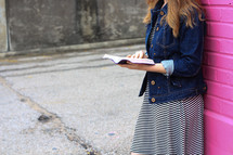 a teen girl in a jean jacket and striped dress reading a Bible in front of a colorful wall 