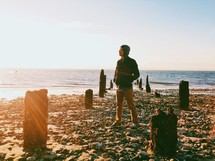 Man standing on the beach between old pier posts looking out the ocean.