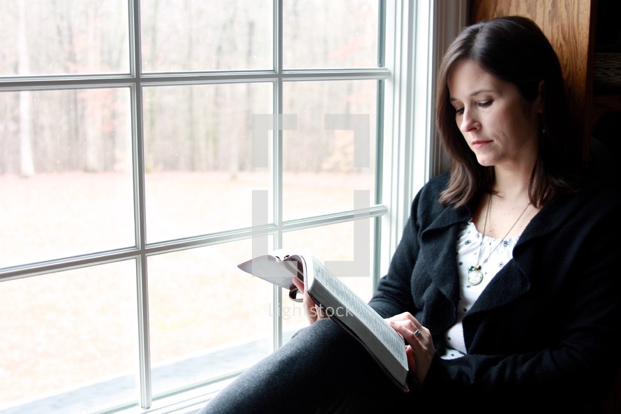 a woman in a sweater sitting in a window reading a Bible 