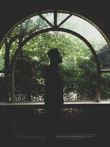 Silhouette of a man standing in front of an arched window over looking trees.