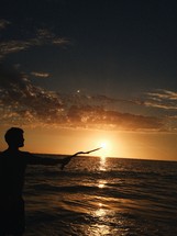 a man with a stick standing on a beach at sunset 
