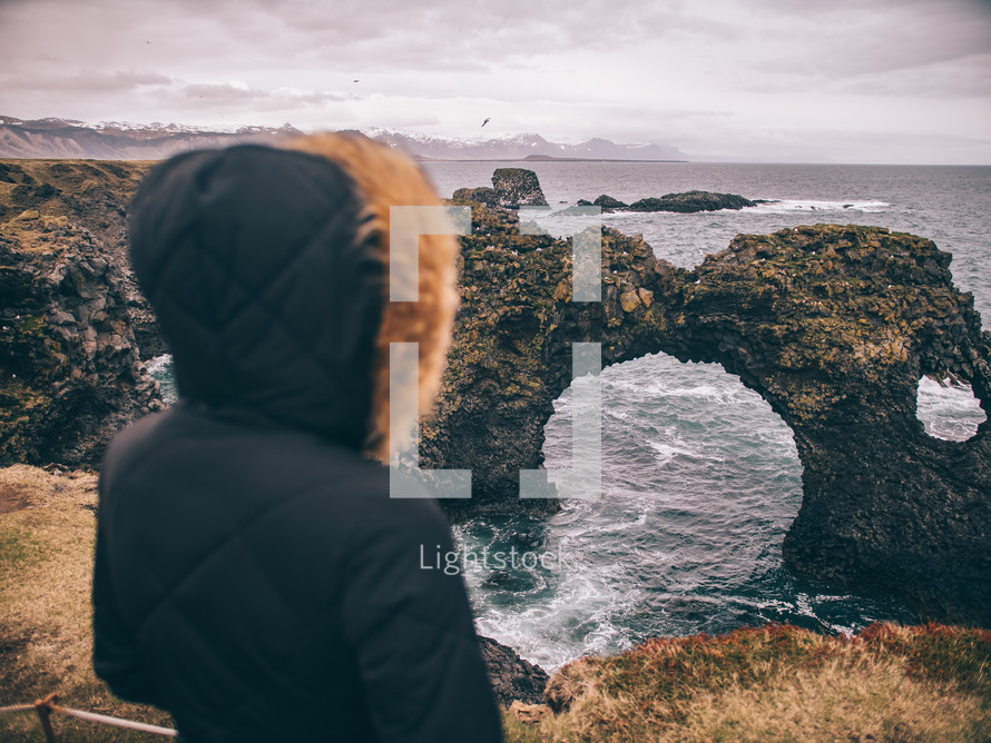 a woman in a coat looking out at a rock arch formation in the ocean 