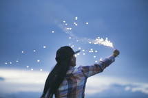 A woman holding a fiery sparkler against the evening sky.