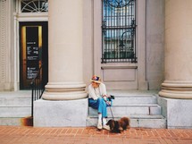Woman sitting on the steps of a building near a pillar, holding her dog's leash.