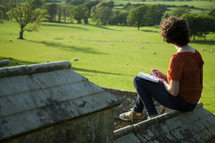 a woman writing in a journal overlooking a flock of sheep 