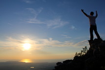 man standing on a mountain top looking out with raised hands  to God  with rising / setting sun in the background