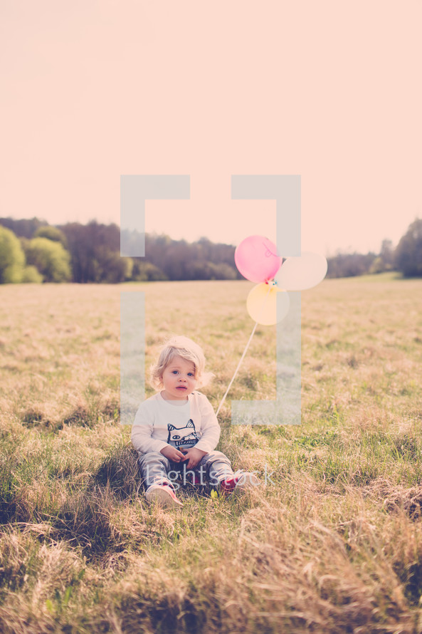 Child sitting in a field of grass holding balloons.