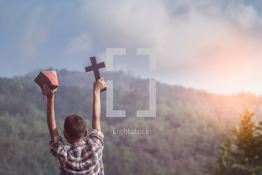 Boy holding holy Bible and christian cross outdoors with raised hands 