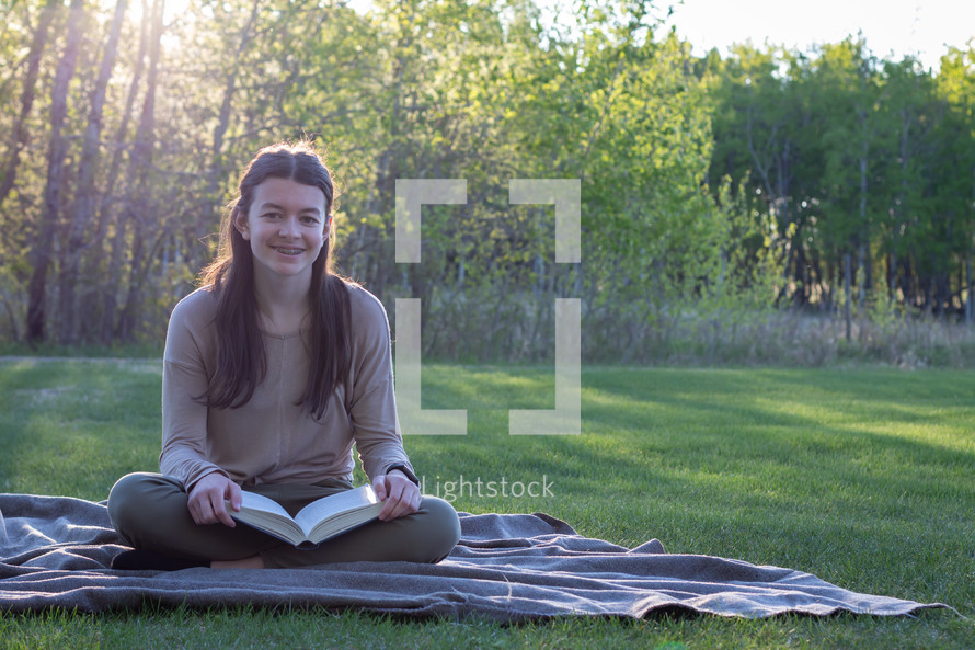 smiling girl reading a Bible sitting on a picnic blanket 