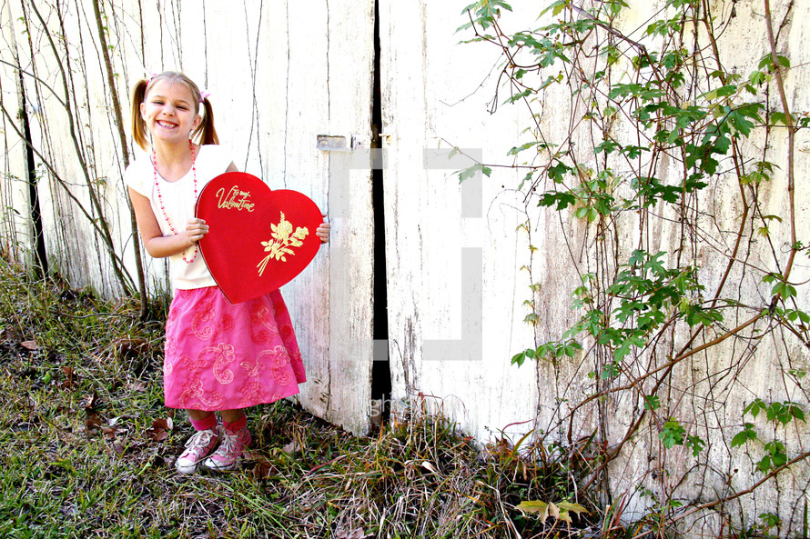 A little girl smiling and holding a big red Valentine box near a white fence.