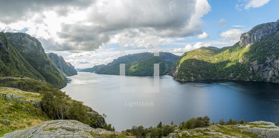 A mountain range in Stavanger, Norway with a lake below and the skies filled with clouds during daytime. In the foreground, we see a rocky landscape. 