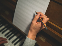 A woman's hands with pencil about to write on music notation paper, while playing a chord on the piano keyboard