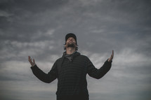 a man standing under a cloudy sky with hands raised 
