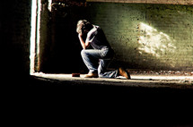 Man in jeans kneeling in prayer in front of sunlit doorway of empty brick room and dirty cement floor.