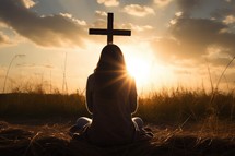 Silhouette of a woman sitting on the grass praying in front of a cross at sunset