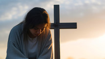 Young woman praying with a cross in the background at sunset