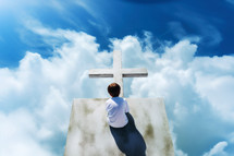 Young boy sitting on top of a cross against blue sky with white clouds
