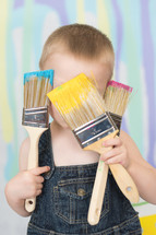 A little boy in overalls covering his face with paint brushes.