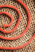 Red rope curled up forming a spiral on a background of light brown jute, with a blurred effect on the bottom