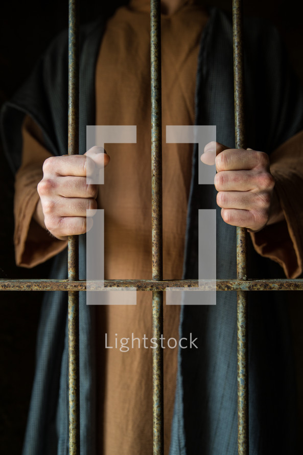 A man in Biblical garb holds onto prison cell bars.