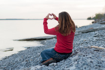 Woman sitting on the beach makes heart hands over the ocean.