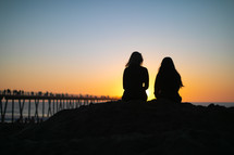 two woman sitting on a beach at sunset 