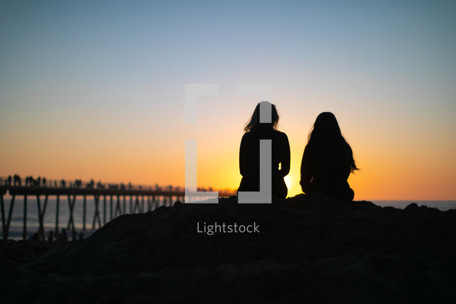 two woman sitting on a beach at sunset 