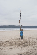 a toddler holding a large stick on a beach in a winter coat 