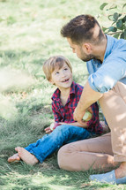 a father and son sitting in the grass holding apples 
