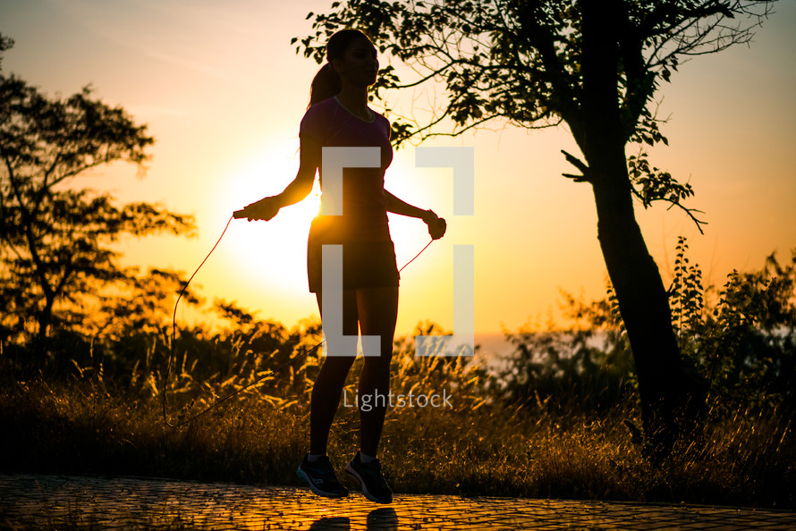 The girl is jumping rope in the park. Young girl doing sports in the forest near sea. Woman is jumping rope in the park. A young girl is doing sports in the forest