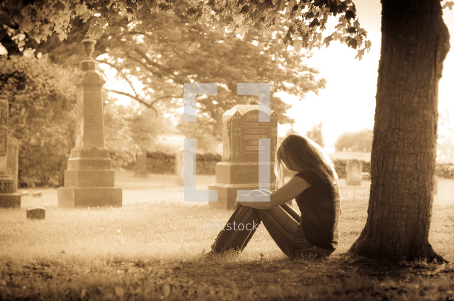 woman sitting in front of a tree in a cemetery grieving 