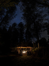People gathered around a fire under a small wood gazebo at night under stars in a forest
