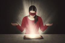 A woman reading a glowing Bible wearing a blindfold. 