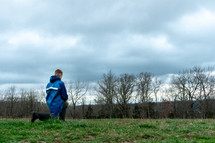 a boy in rain boots kneeling looking out a forest 