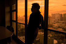 a man standing in a window with view of a city behind him 