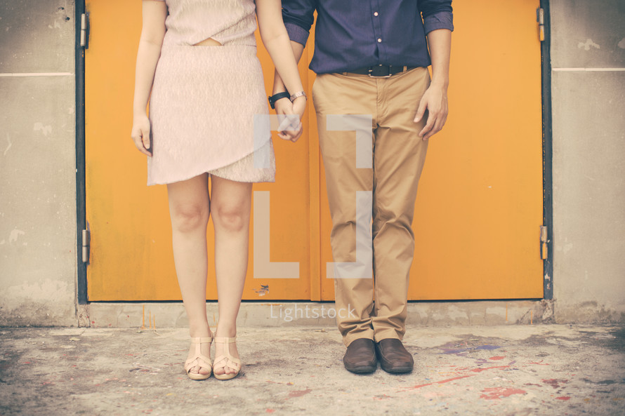 legs of a couple holding hands standing in front of yellow doors 