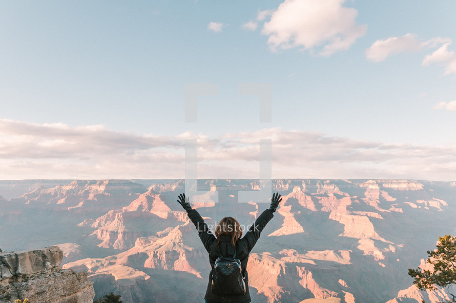 a woman standing at the edge of a cliff in front of canyons with arms raised 