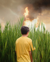 A young boy is standing alone outside in a grass field looking at a burning fire in the distance for a trauma, anxiety metaphore or danger message