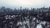 Aerial shot of big park in downtown Toronto on a snowy winter day. Drone flying over leafless trees while showing urban cityscape. Modern high-rises in the distance. Aircraft moving right sideward.